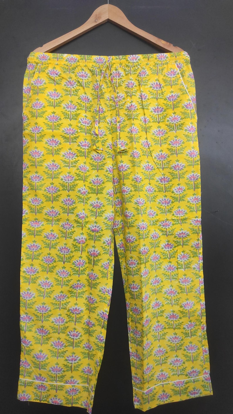 Op de afbeelding: Gele pyjamabroek met een bloemenpatroon in roze, groen en wit. De broek hangt aan een houten kleerhanger. Het bloemenpatroon herhaalt zich over de stof.