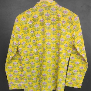 Op de afbeelding: Een gele pyjama-top met lange mouwen en een bloemenpatroon van roze en groene bloemen. De pyjama-top hangt aan een houten hanger tegen een donkere achtergrond. Het bloemenpatroon wordt over de hele stof herhaald.