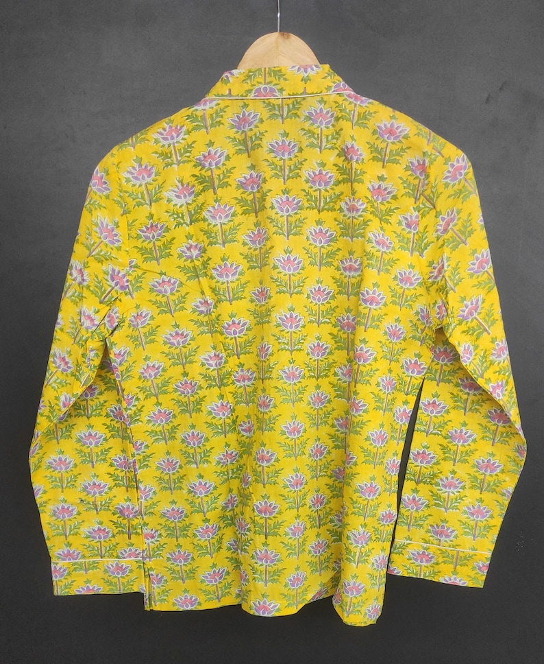 Op de afbeelding: Een gele pyjama-top met lange mouwen en een bloemenpatroon van roze en groene bloemen. De pyjama-top hangt aan een houten hanger tegen een donkere achtergrond. Het bloemenpatroon wordt over de hele stof herhaald.