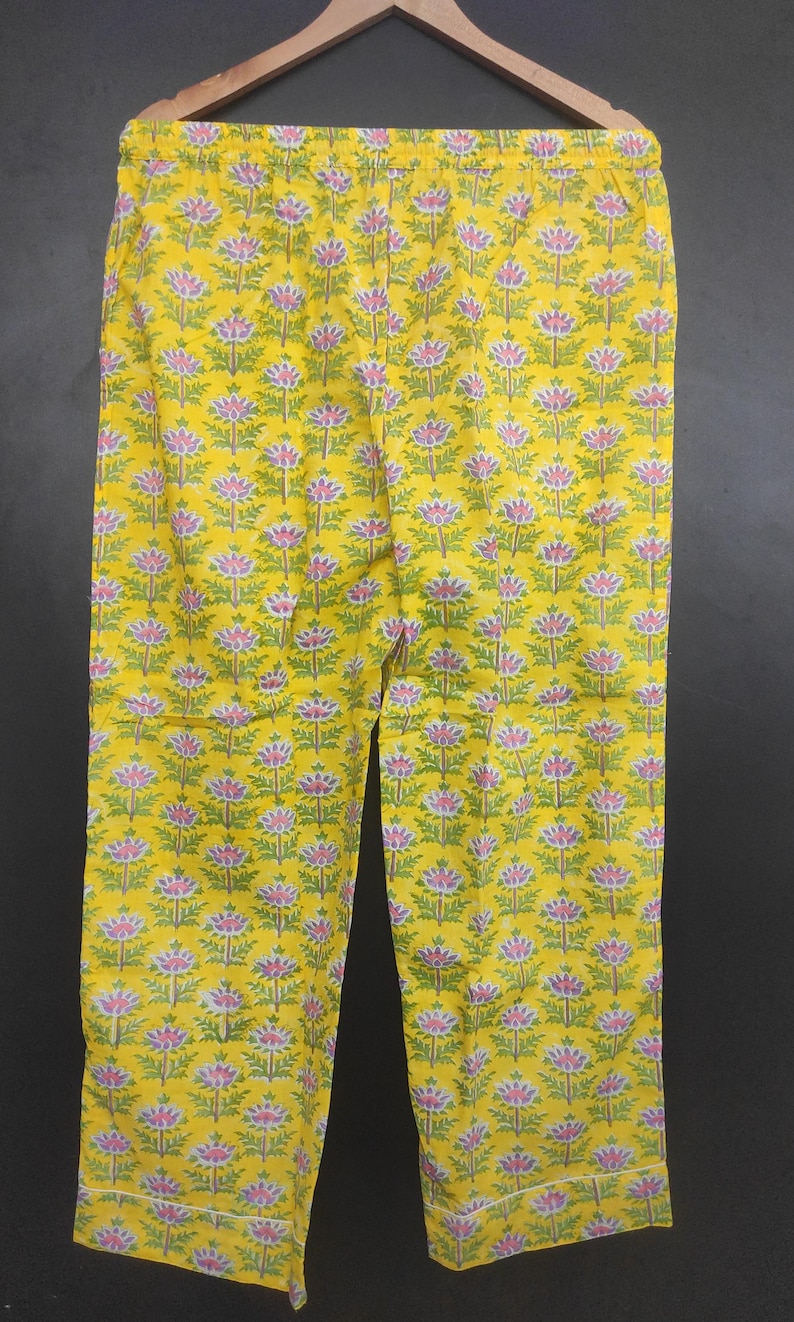 Op de afbeelding: Gele pyjamabroek met een herhalend bloemenpatroon in roze, groen en wit. De broek is gemaakt van een lichte stof en heeft een comfortabele, relaxte pasvorm. Het bloemmotief is gelijkmatig verdeeld over het gehele oppervlak.