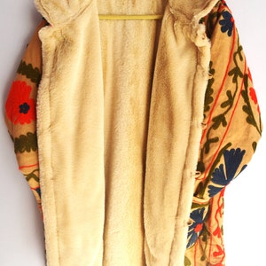 May include: A tan jacket with a floral embroidered design in shades of red, green, blue, and orange. The jacket is lined with a soft, cream-colored faux fur.