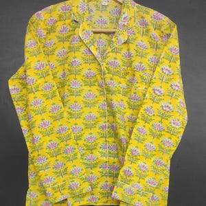 Op de afbeelding: Gele pyjama set met een bloemenpatroon in roze, groen en wit. De top met lange mouwen heeft een reverskraag, een borstzak en witte biezen. De pyjama hangt aan een houten hanger.