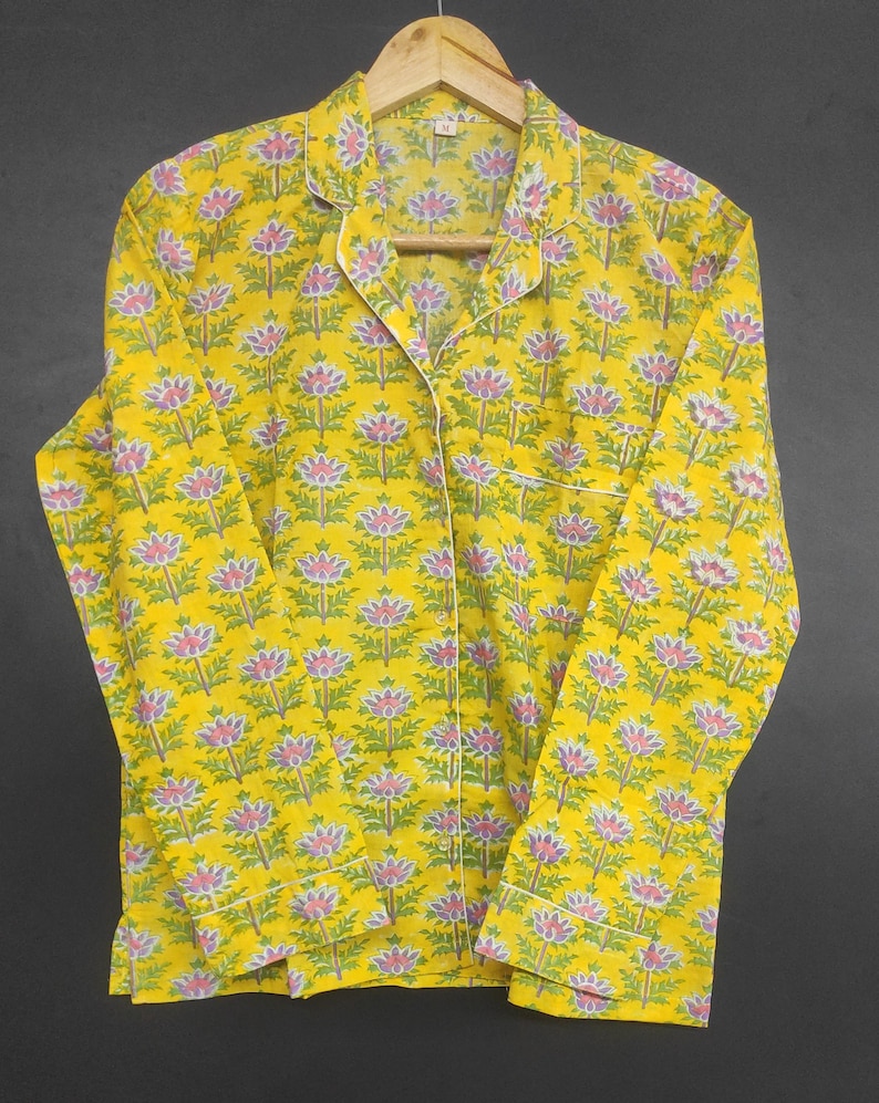 Op de afbeelding: Gele pyjama set met een bloemenpatroon in roze, groen en wit. De top met lange mouwen heeft een reverskraag, een borstzak en witte biezen. De pyjama hangt aan een houten hanger.