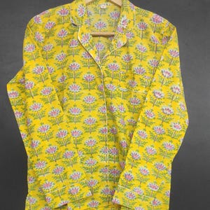 Op de afbeelding: Gele pyjama set met bloemenpatroon. De top met lange mouwen en de broek hebben een herhalend ontwerp van roze en paarse bloemen met groene bladeren op een gele achtergrond. Witte biezen accentueren de kraag, manchetten en zak.
