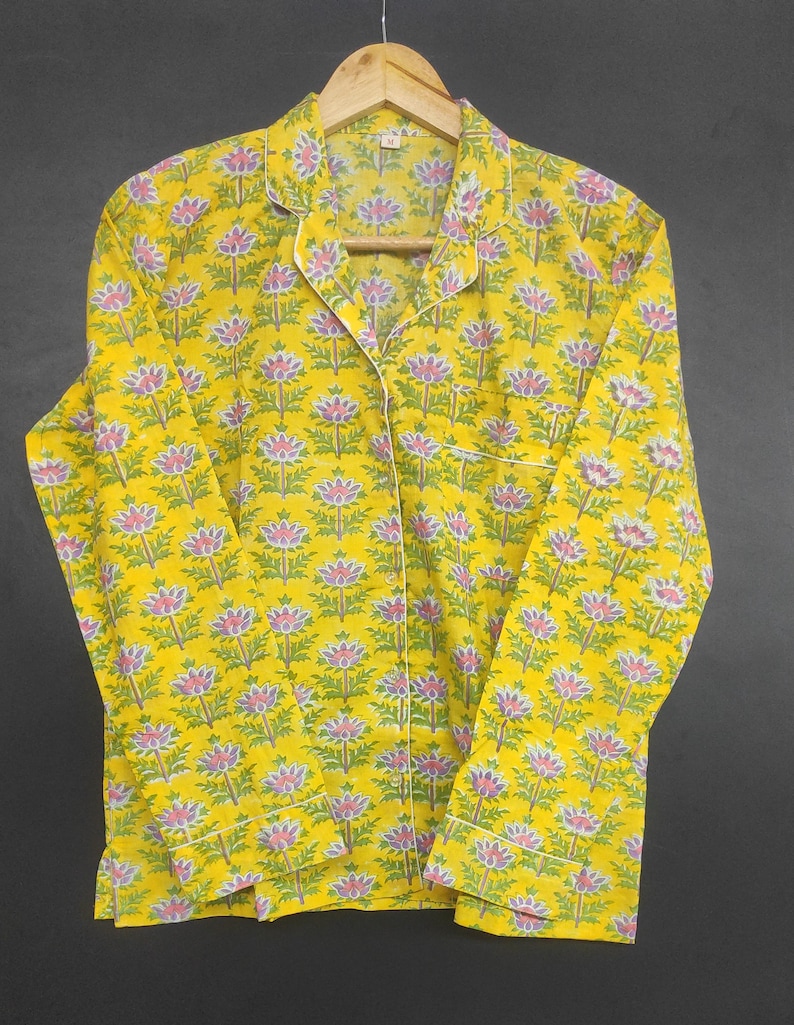 Op de afbeelding: Gele pyjama set met bloemenpatroon. De top met lange mouwen en de broek hebben een herhalend ontwerp van roze en paarse bloemen met groene bladeren op een gele achtergrond. Witte biezen accentueren de kraag, manchetten en zak.