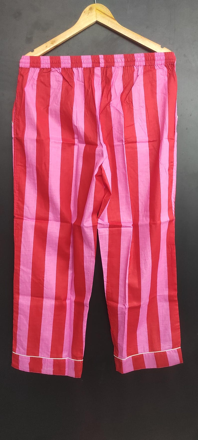 K&ouml;nnte beinhalten: Rote und rosa gestreifte Schlafanzughose mit wei&szlig;em Saum. Die Hose h&auml;ngt an einem Holzb&uuml;gel.
