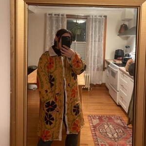 May include: A long, yellow, floral patterned jacket with a white fur collar. The jacket has a button closure and is embroidered with black, orange, and green flowers.