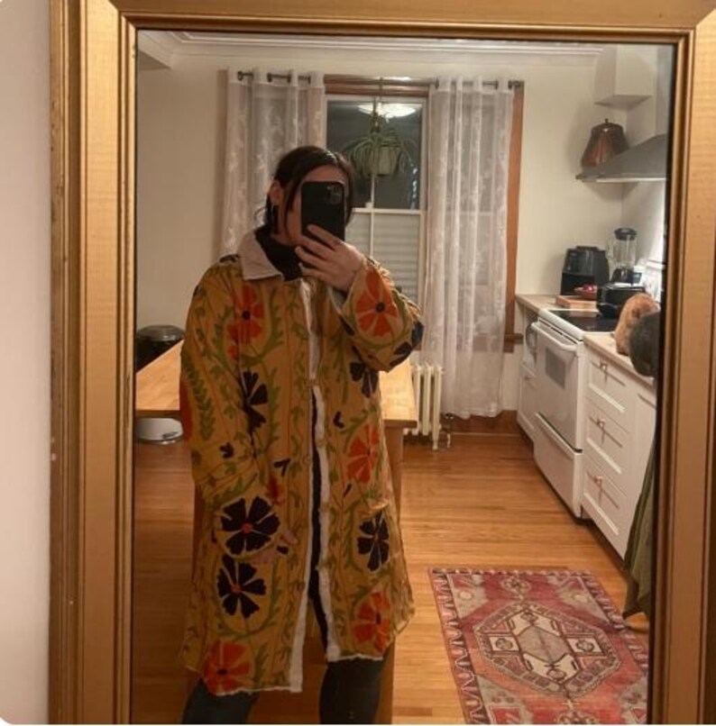 May include: A long, yellow, floral patterned jacket with a white fur collar. The jacket has a button closure and is embroidered with black, orange, and green flowers.