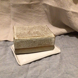 Könnte beinhalten: Eine weiße Keramik-Butterdose mit gesprenkeltem Glase und einem braunen Band um den oberen Rand. Die Dose steht auf einem weißen Keramiktablett.