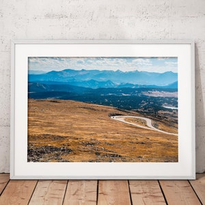 May include: A framed photograph of a winding road leading through a mountainous landscape. The road is surrounded by brown grass and the mountains are a hazy blue in the distance.
