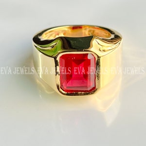 Puede incluir: Un anillo de oro con una piedra preciosa roja cuadrada grande. El anillo tiene una banda gruesa y un diseño simple.