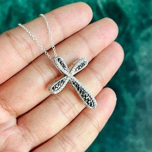 May include: A silver cross pendant necklace with black and white diamonds. The cross is a simple design with a slightly curved shape.