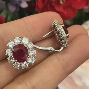 May include: A pair of silver-toned earrings featuring a central oval-cut deep red gemstone surrounded by a halo of twelve clear round-cut stones. The earrings have a hinged lever-back closure mechanism and a metallic setting with a polished finish.