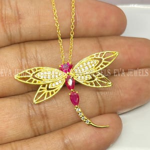 May include: A gold-colored dragonfly pendant necklace. The dragonfly has openwork wings, a body of red gemstones, and a tail of small white gemstones. The wings are embellished with small white gemstones. The necklace is on a gold chain.