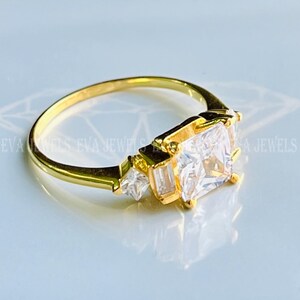 May include: A gold ring featuring a large square-shaped clear stone and two smaller clear stones on either side.