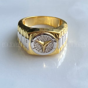 May include: A gold and silver ring with a circular design featuring a Y-shaped symbol surrounded by small clear stones.