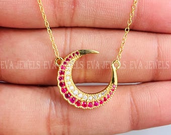 May include: A gold chain necklace with a crescent moon pendant. The pendant is adorned with small red and white stones.