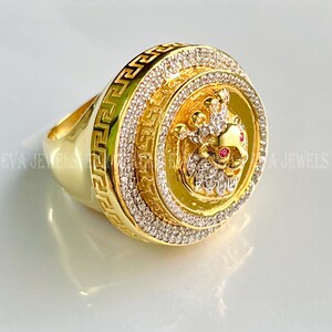 Könnte beinhalten: Ein goldener Ring mit einer großen, runden Fläche, die einen Löwenkopf mit Krone und roten Rubinaugen zeigt. Der Ring ist mit einem griechischen Schlüsselmuster und klaren Zirkonia-Steinen verziert.