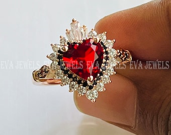 Könnte beinhalten: Ein Ring aus Roségold mit einem großen roten herzförmigen Edelstein, umgeben von einem Heiligenschein aus kleineren weißen und schwarzen Edelsteinen. Der Ring hat eine zarte Band und ein kronenartiges Design.