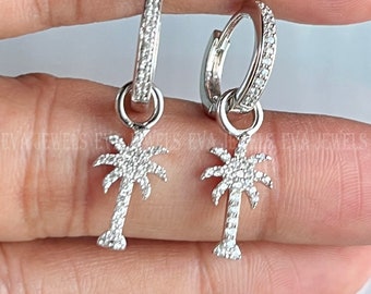 May include: A pair of silver hoop earrings with a dangling palm tree charm. Each charm is covered in small, clear cubic zirconia stones.