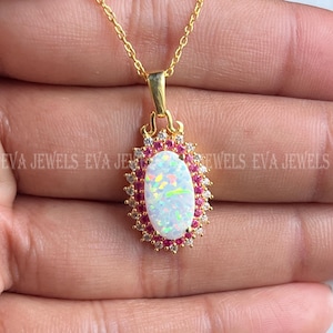 Puede incluir: Un collar dorado con un colgante ovalado de ópalo. El ópalo está rodeado de pequeñas piedras preciosas rosas y transparentes. El colgante cuelga de una delicada cadena dorada. El texto "EVA JEWELS" es visible en el fondo.