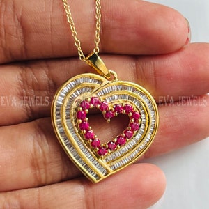 May include: A gold heart-shaped pendant necklace with a smaller heart-shaped cutout in the center. The pendant is adorned with clear baguette-shaped stones and red round stones.