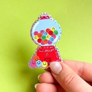 Puede incluir: Una colorida pegatina de máquina de chicles con una base roja y una parte superior azul llena de chicles con caras sonrientes. La pegatina tiene detalles de purpurina sobre un fondo verde brillante.