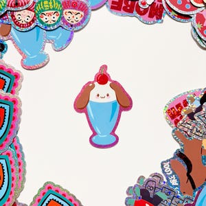 May include: A collection of colorful, glittery stickers on a white background. The stickers feature various designs, including a sundae with a cherry on top, Japanese dolls, and heart shapes. The stickers have a holographic effect and are outlined in pink.