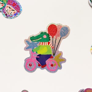 Puede incluir: Una pegatina brillante con un cocodrilo verde montando en bicicleta rosa y sosteniendo globos. El cocodrilo lleva un suéter verde, pantalones azules y una bufanda a rayas rojas y blancas. Los globos son rojos y azules.
