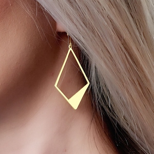 Op de afbeelding: Een vrouw draagt een blauw shirt en een gouden ketting met een geometrische gouden oorbel.