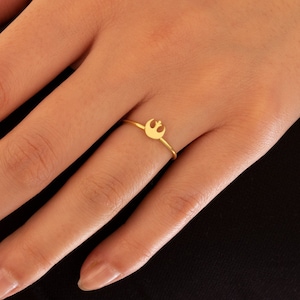 May include: A gold ring with a small Rebel Alliance symbol. The ring is worn on a person's finger.