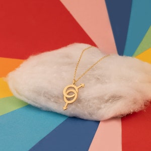 May include: A gold necklace with a pendant featuring two interlocking circles and an arrow. The necklace is displayed on a white fluffy surface against a colorful rainbow background.