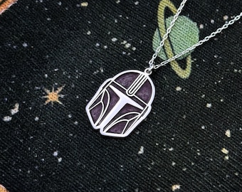 May include: A silver necklace featuring a pendant shaped like a Mandalorian helmet. The helmet has a dark purple background with white and silver details. The necklace is displayed on a black fabric with a space-themed design.