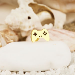 May include: A gold-colored video game controller charm. The controller is a simple design with a cross, a circle, and two buttons on the front.