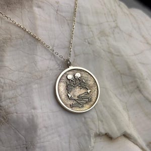 May include: Silver-toned pendant necklace featuring a round charm with an embossed design of two skeletons embracing. The pendant hangs from a delicate chain. The background is a light, textured surface.