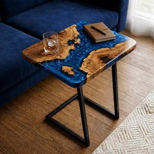 May include: A rectangular side table featuring a natural live-edge wood slab with a vibrant blue epoxy resin river inlay. The table is supported by a black metal frame with a C-shaped base, holding a glass of liquid and a brown notebook.