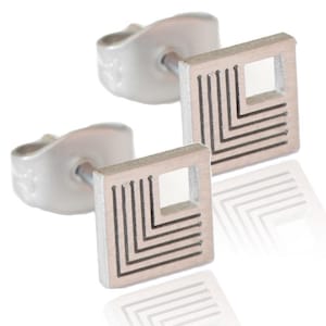Könnte beinhalten: Ein Paar silberne Ohrstecker mit geometrischem Design. Die Ohrringe haben eine quadratische Form mit einem kleineren quadratischen Ausschnitt in der Mitte. Das Design besteht aus dünnen Linien, die einen geschichteten Effekt erzeugen.