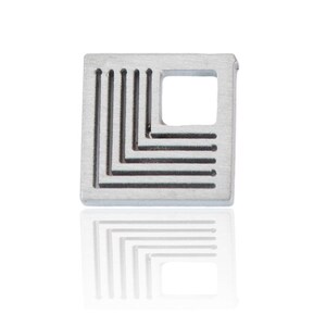 Könnte beinhalten: Ein quadratischer Anhänger aus silbernem Metall mit einem geometrischen Muster aus Linien und einem kleineren quadratischen Ausschnitt in der Mitte.