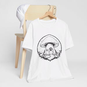 May include: White t-shirt with a black and white illustration of a mushroom house with a door and windows. The mushroom is surrounded by smaller mushrooms.