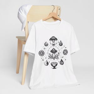 May include: White t-shirt with a black and white graphic design of a mushroom with a circle of symbols around it. The design includes a mushroom, a chalice, and other symbols.