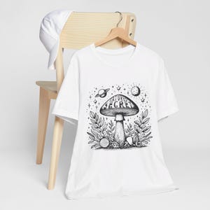 May include: White t-shirt featuring a black and white illustration of a mushroom with the text "Nature's Secret" on the cap. The mushroom is surrounded by leaves and stars.
