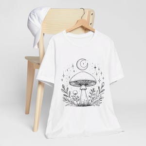 May include: White t-shirt with a black and white illustration of a mushroom, crescent moon, and stars. The design is centered on the shirt and surrounded by leaves and other small mushrooms. The shirt is hanging on a wooden hanger.