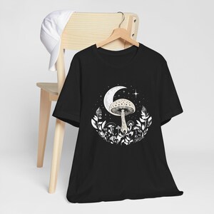 May include: Black t-shirt with a white graphic of a mushroom under a crescent moon and stars. The mushroom is surrounded by leaves and smaller mushrooms.