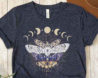 May include: A dark heather grey t-shirt with a graphic print of a moth with a skull and crossbones design. The moth is surrounded by a crescent moon and stars.