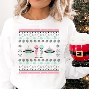 May include: White Christmas sweater with a pink and green alien design. The aliens are wearing spacesuits and standing in front of flying saucers. The sweater has a snowflake pattern and a pink and white border.