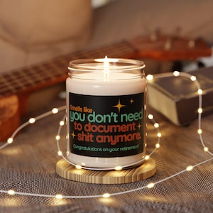 May include: A lit candle in a glass jar with a black label that reads "Smells like you don't need to document shit anymore. Congratulations on your retirement!" The candle sits on a round wooden coaster, with string lights and a guitar in the background.