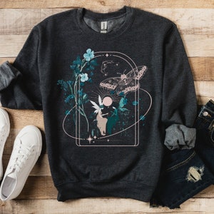 May include: Dark gray sweatshirt featuring a whimsical design with a moth, flowers, and a fairy-like figure within a rectangular frame. The design incorporates teal, pink, and white elements. The sweatshirt is paired with white sneakers and blue jeans.