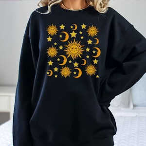 May include: Black sweatshirt with a graphic print of yellow suns and moons with stars on a black background.