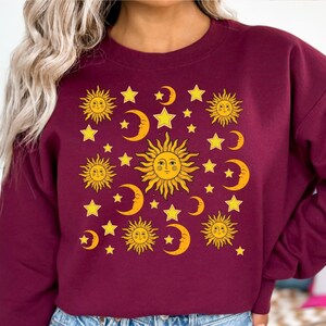 May include: A maroon sweatshirt with a graphic print of yellow suns, moons, and stars. The graphic is a repeating pattern of suns with faces, crescent moons, and five-pointed stars.