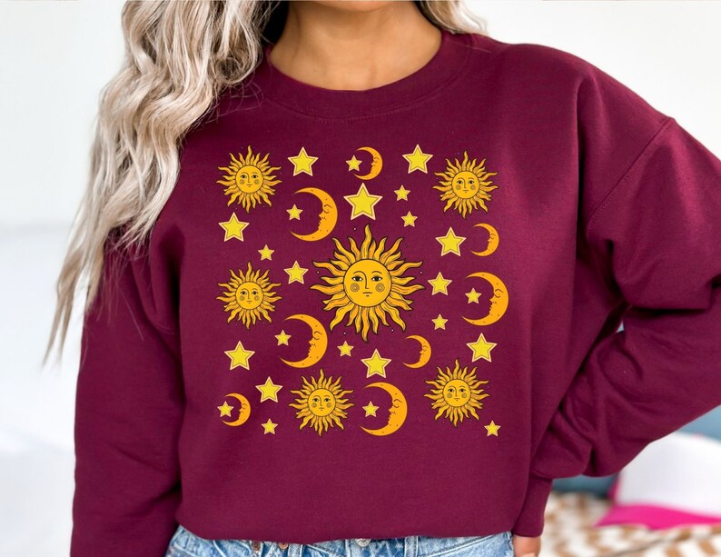 May include: A maroon sweatshirt with a graphic print of yellow suns, moons, and stars. The graphic is a repeating pattern of suns with faces, crescent moons, and five-pointed stars.
